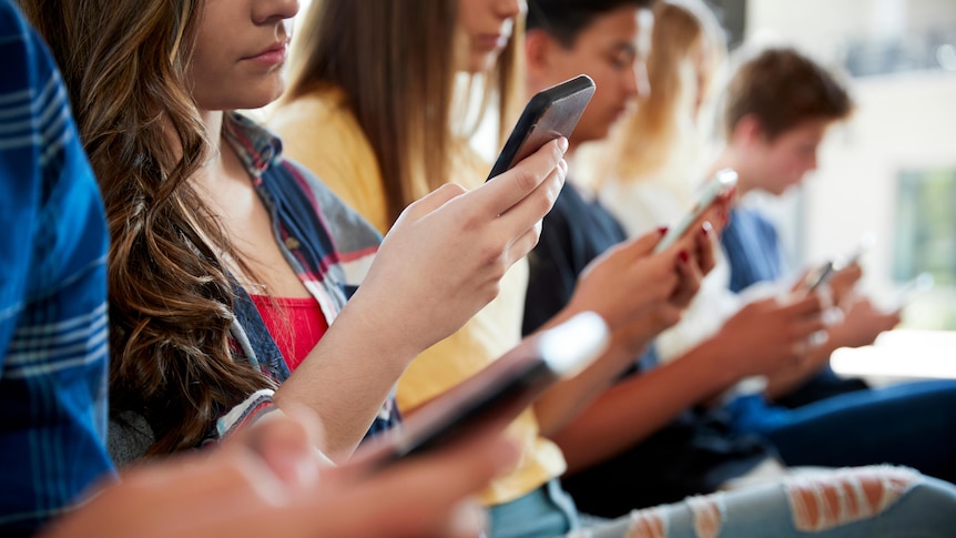 A row of teenagers sitting down on a long bench chair, all looking at their smartphones