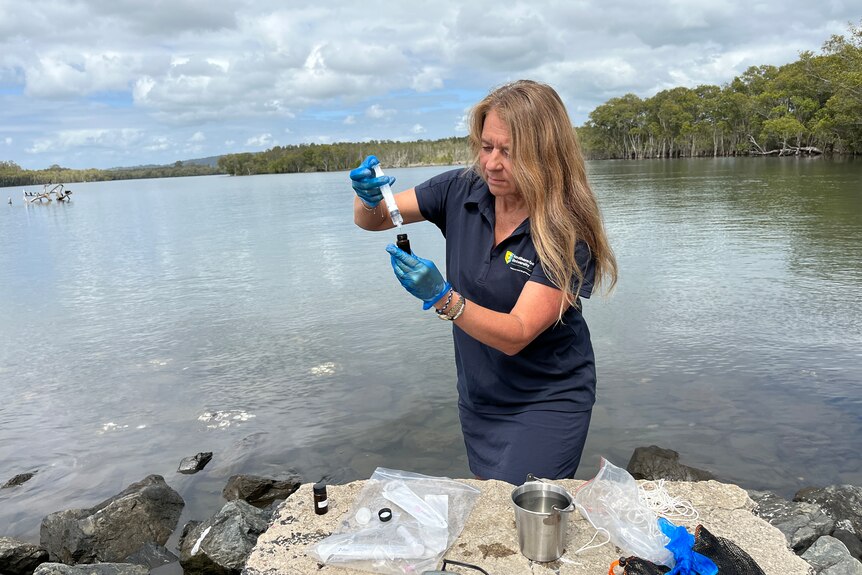 A woman stands on the edge of a river, taking a water sample.