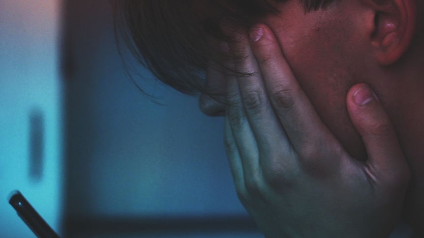 A close up image of a male teen with his hand over his face looking at a mobile phone