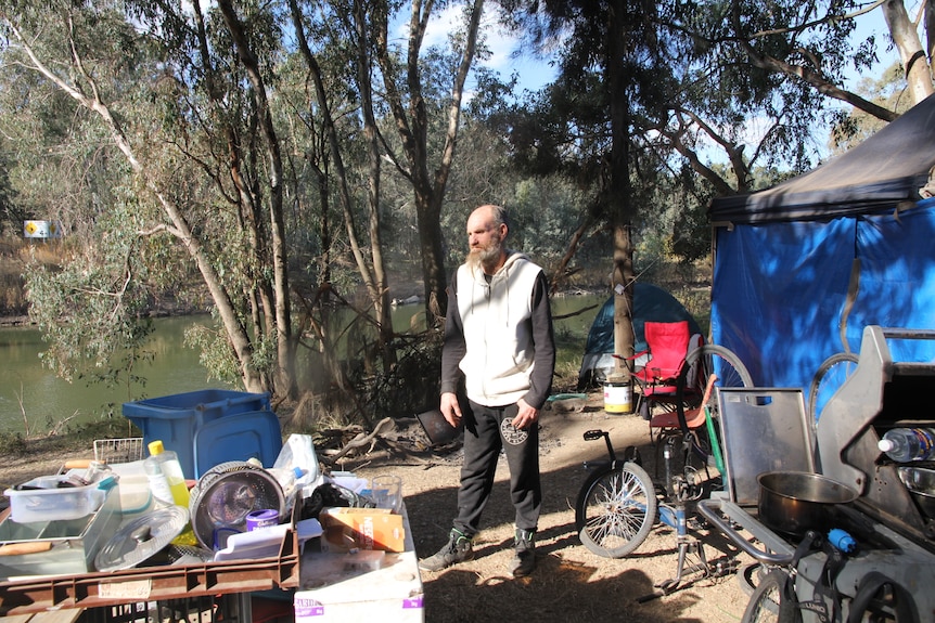 A bearded man stands on the banks of a river at his makeshift home made of tents and tarps