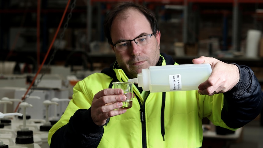 A bespectacled man with dark hair carefully pours liquid from a plastic bottle into a small beaker.