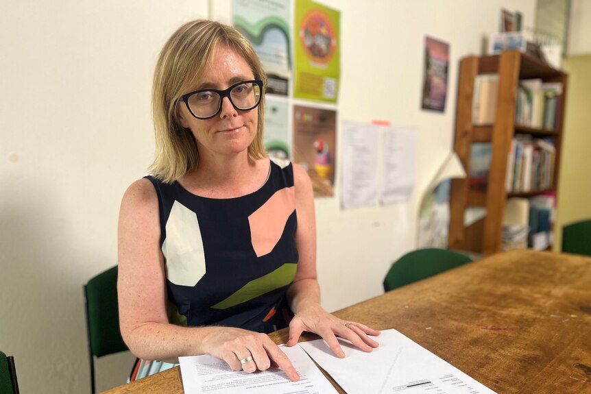 Woman with glasses sits at table at points to documents on it