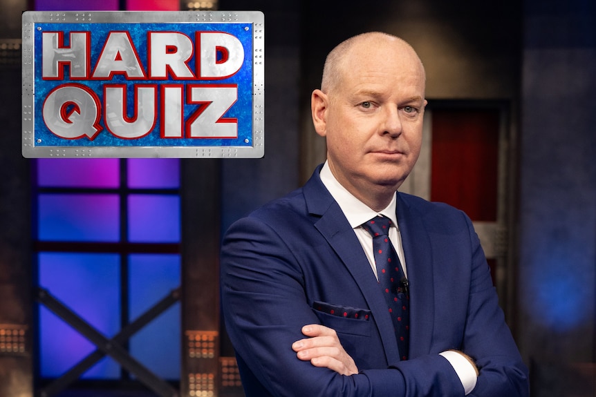 Tom Gleeson standing with his arms crossed with a serious look on his face with th Hard quiz logo in the top left corner