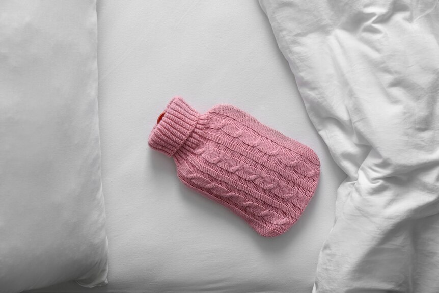 A hot water bottle in a pink knitted cover lies on white sheets in a bed.