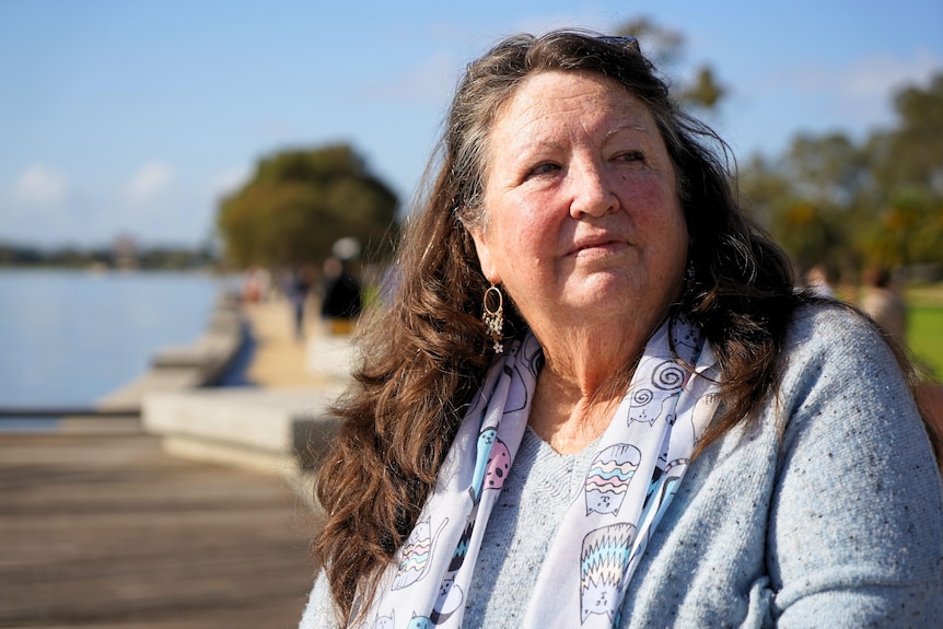 A woman with brown hair and a grey jumper, sits in the sun by a river.