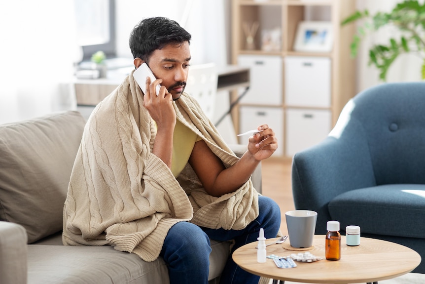 A man sits on a couch, draped in a blanket, checks his temperature, while talking on a smartphone.