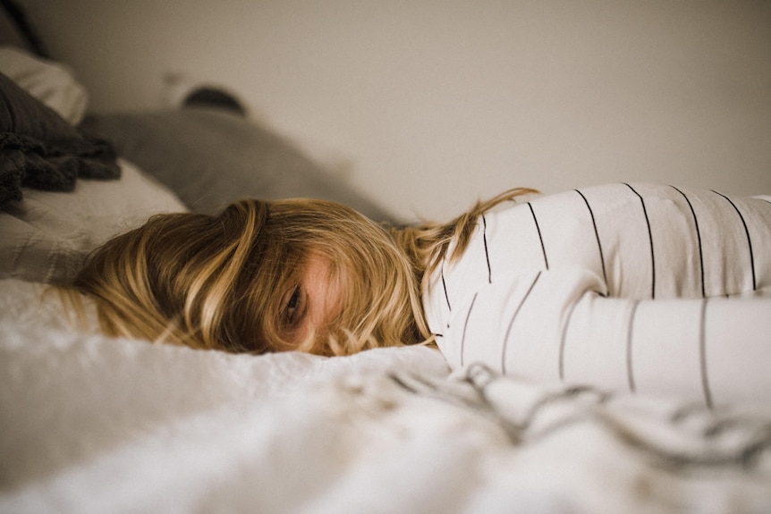 A blonde woman in a white shirt lies in bed and faces the camera, her eyes visibile through her hair.