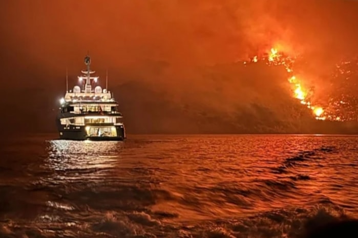 A yacht sits offshore of a burning forest, at night.