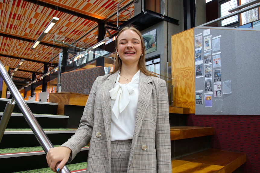 A young woman wearing a grey blazer stands on a stairwell, smiling.