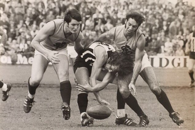 A photo showing a man playing football while being tackled by two opponents.