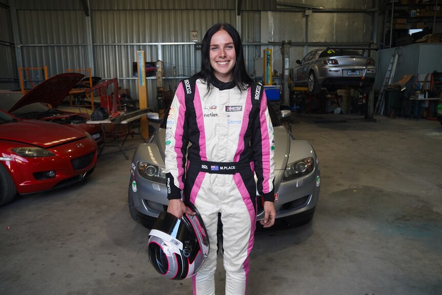 A young woman wearing motorsport gear and standing in front of a car.