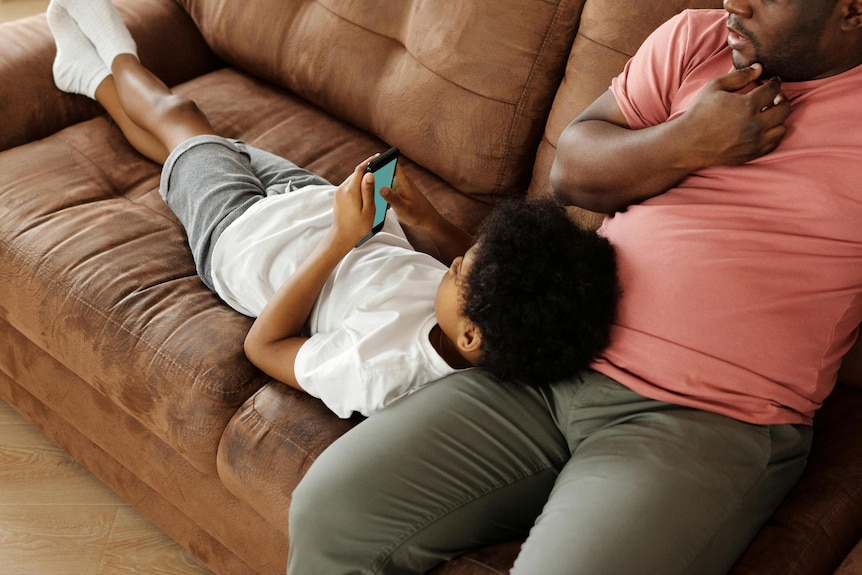 son lays on dad's lap on the couch looking at phone