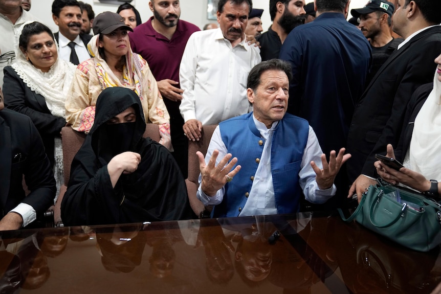 Bushra Bibi sits next to Imran Khan who are both behind a table surrounded by people at a press conference