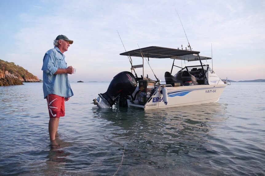 Man stands in front of a boat, ankle-deep in ocean water, looking pensively out to sea.