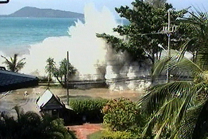 A video still of a tsunami wave coming over a wall and sending up a big spray of water. The blue ocean is in the background