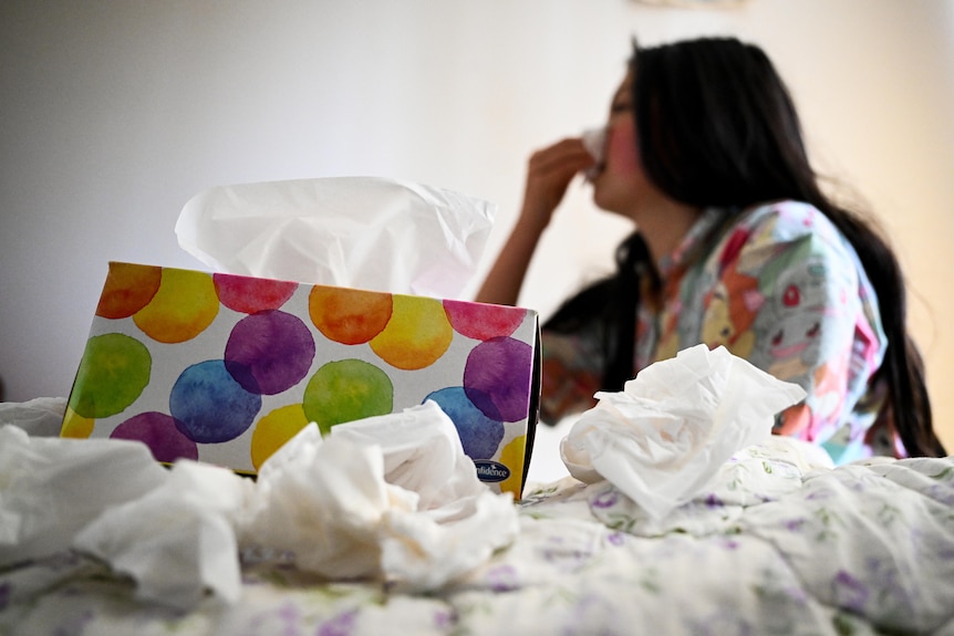 A woman blows her nose with a box of tissues in front of her