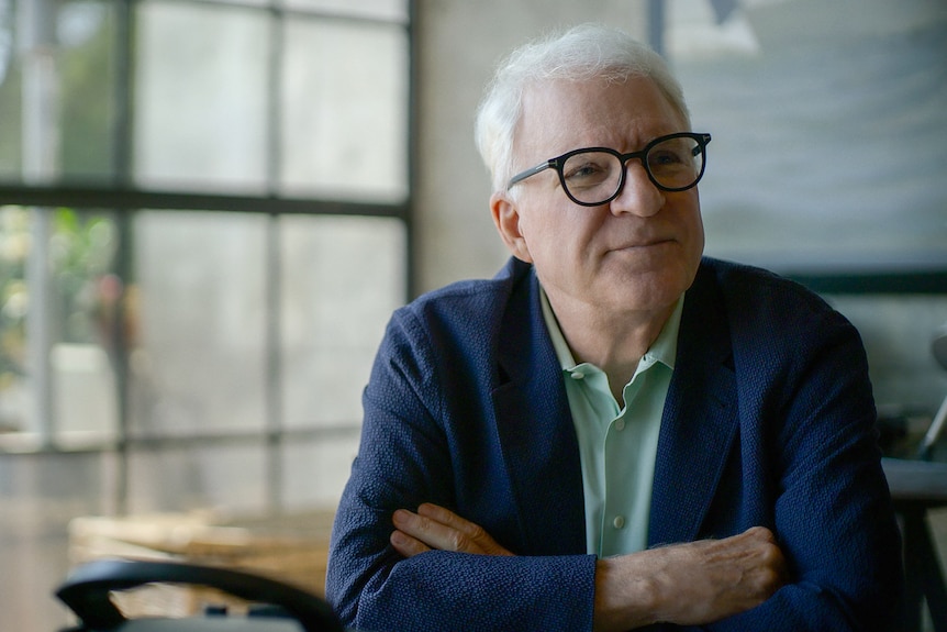 Steve Martin looks slightly to the right as he sits at a table with his arms crossed in front of him, a pensive look on his face