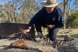 A woman in a broad tan hat bends down while making a fire.