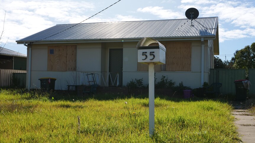 An abandoned house with boarded up windows and long grass.