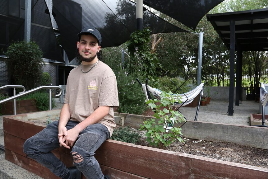 A young man in ripped jeans and a hat sits next to a garden