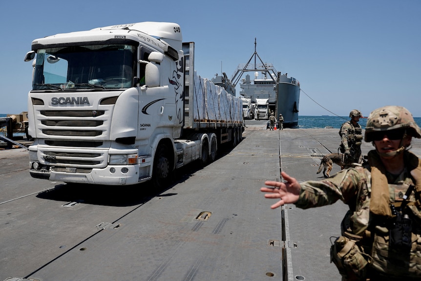 A soldier motions to the camera to move away as a huge truck drives into frame onto a pier off a huge boat