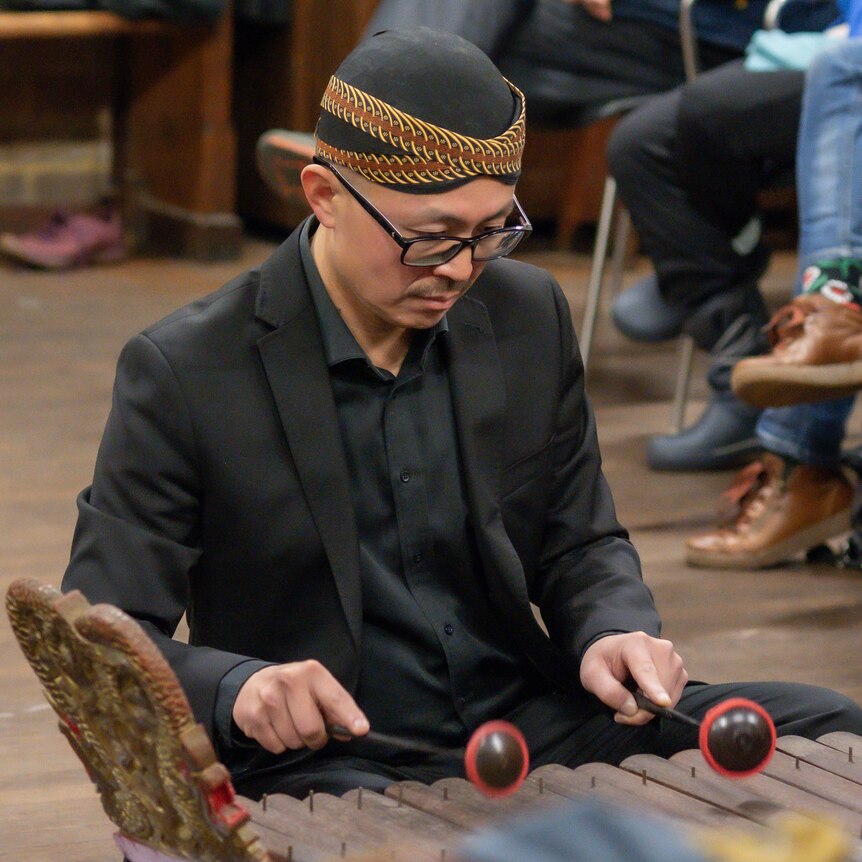 A man in his early 40s playing a wooden-keyed instruments. He's holding two mallets while being seated on the floor.