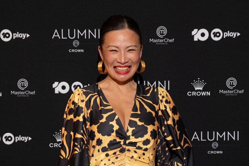 An Asian woman with black hair in a gold and black dress smiles in front of a black background