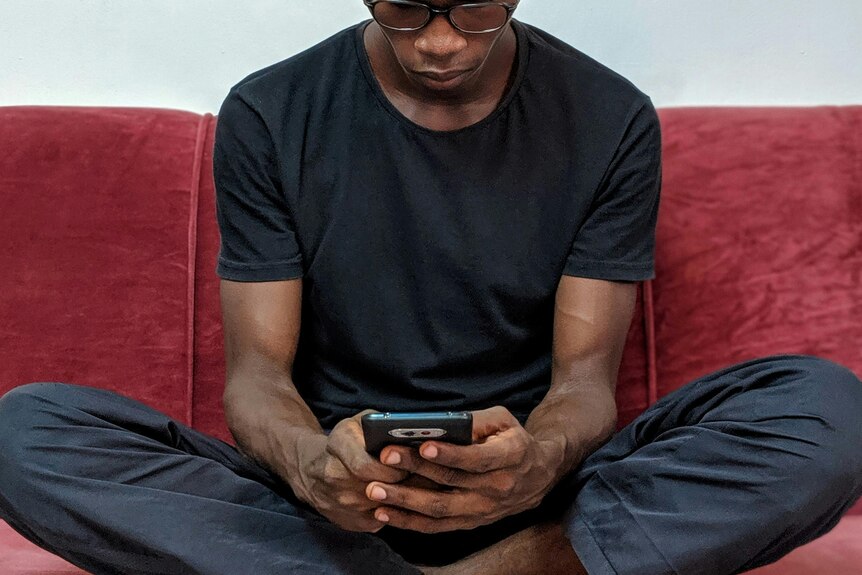 man sits cross-legged on couch looking down at phone