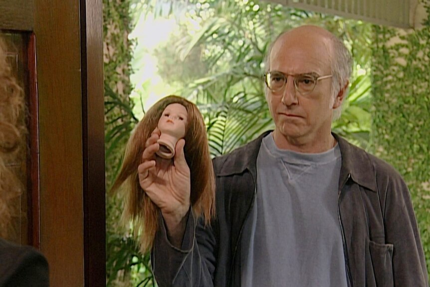 Larry stands in a doorway and holds the head of a brunette doll up with a serious expression on his face.