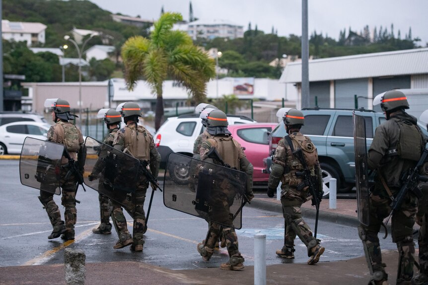 Troops in uniform walk away from the camera holding riot shields in the street