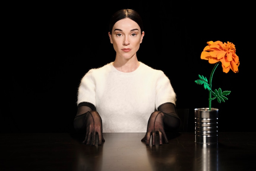 St Vincent sits in darkness, wearing white jumper and black gloves, at a table with an orange flower in a tin can