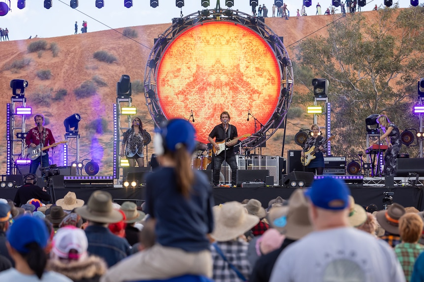 Man plays guitar on stage with large circular orange light behind him.