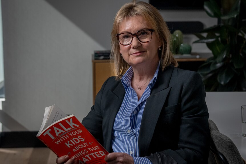 An older woman with a blonde bob, black blazer and blue shirt holding a red book.