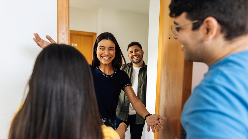 Young couple welcoming male and female guests at an apartment doorway, with smiles and arms outstretched. 