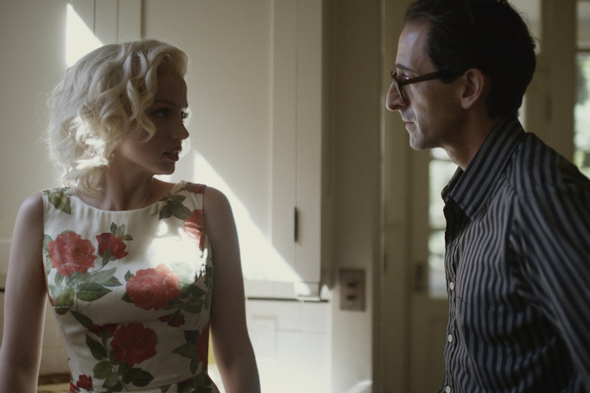Blonde white woman in floral 50s dress looks at white man with dark hair and round glasses in navy striped shirt