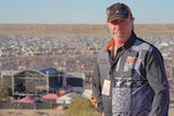 A man in a cap stands on a sandune hill with a big festival crowd behind him