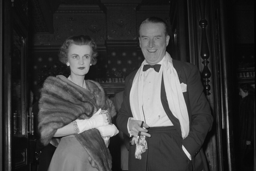 Margaret Campbell dressed in a fur coat clutches a purse standing next to a man dressed in a suit with a bowtie.