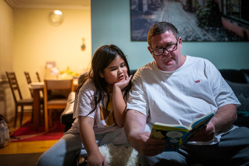 A man in glasses and a white t-shirt holds a book out as a younger girl reads aloud and leans into him in their living room.