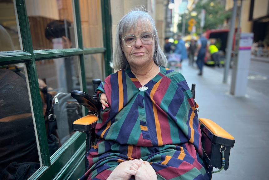 A middle aged white woman without a right arm sits outside in a motorised wheelchair, wearing a striped shirt and big glasses. 