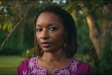 A close-up of a middle-aged Black woman wearing a purple blouse looks directly into the camera. She is standing outside.