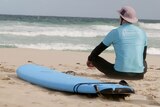 A man sitting on the sand looking out at the ocean with a surfboard next to him 