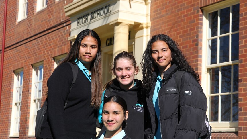 four girls standing together smiling, outside with a brick building behind them