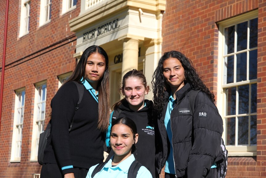 four girls standing together smiling, outside with a brick building behind them