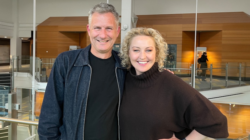 Adam Hills and Zan Rowe in the ABC Melbourne foyer. Zan wears a black jumper and Adam wears a black tshirt and denim jacket