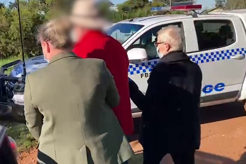 A man in a red shirt being arrested in front of a police car