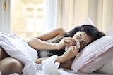 A woman lying on her side curled up in bed during the day, blowing her nose with a tissue.