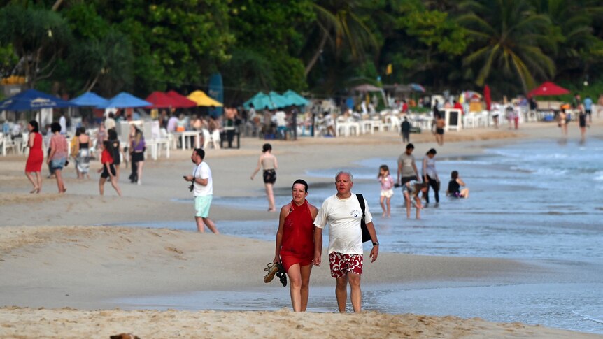 A man and a woman walk along a yellow sand beach and colourful umbrellas dot the shore.