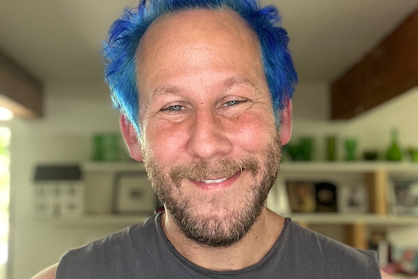 A man with blue hair and brown beard smiles in a close up of his face.