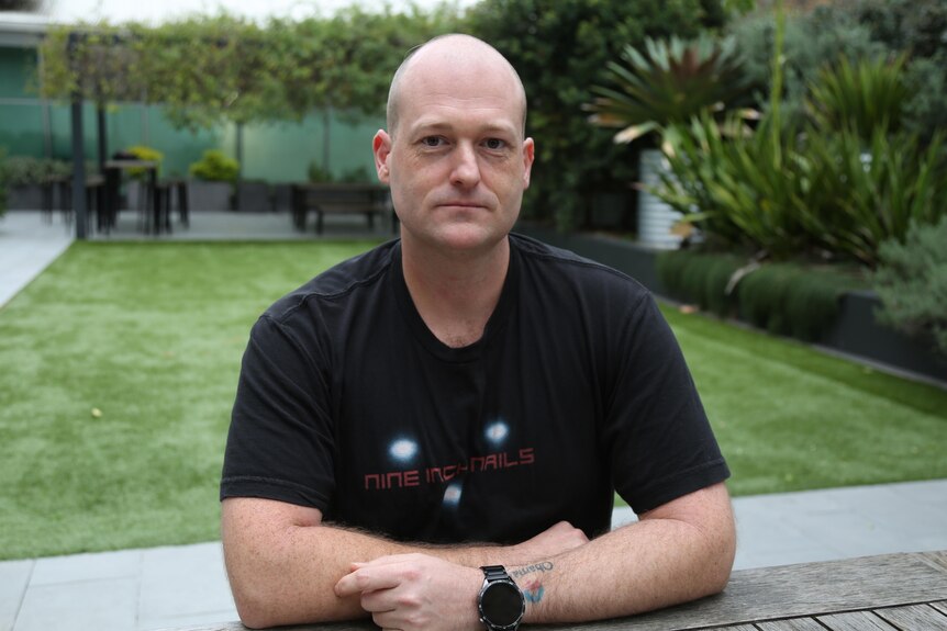 Joel Hill, with bald head and black t-shirt, smiles sitting at wooden table with grass and green bushes behind him.