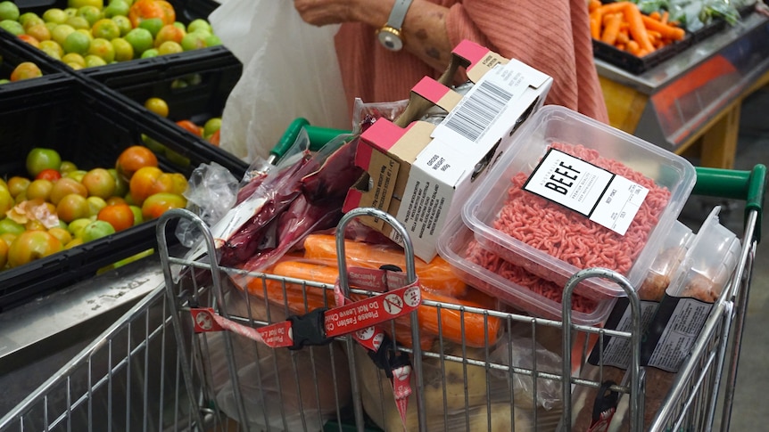 A close-up shot of a shopping trolley full of groceries in a supermarket with a woman behind bagging apples.